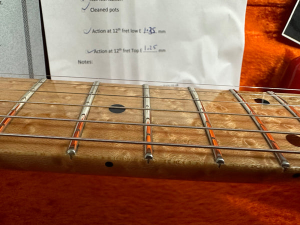 Close-up of guitar fretboard with metal frets and strings, showing birdseye maple wood grain and black dot inlays, with guitar setup notes in background and orange case lining beneath.