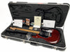Ernie Ball Music Man electric guitar in cherry red finish with maple fretboard, displayed inside a black plush-lined hard case with papers, certificate of authenticity, and accessories.