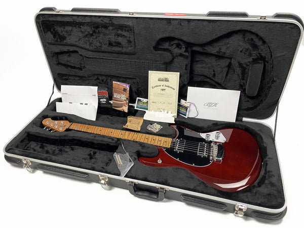 Ernie Ball Music Man electric guitar in cherry red finish with maple fretboard, displayed inside a black plush-lined hard case with papers, certificate of authenticity, and accessories.