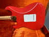Red electric guitar body with white backplate and gold neck plate, resting in orange velvet-lined guitar case