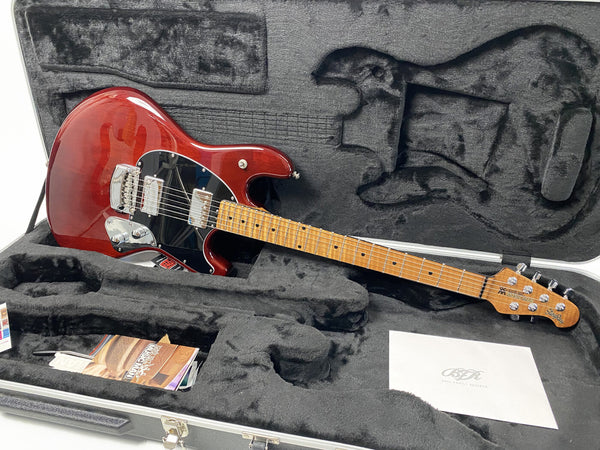 Ernie Ball Music Man StingRay electric guitar in transparent red finish with maple neck and black pickguard, displayed in a black plush-lined guitar case with accessories and paperwork.