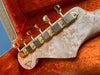 Close-up of guitar headstock with gold tuning pegs and quilted maple wood grain on orange plush guitar case lining