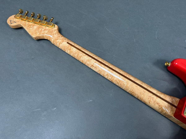 Close-up of guitar neck and headstock with birdseye maple wood and gold tuning pegs, showing back of red electric guitar body against gray background.