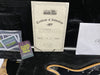 Ernie Ball Music Man Certificate of Authenticity for Ball Family Reserve guitar with serial number 686265 and signed by Scott Ball, displayed inside a guitar case with Ernie Ball guitar string packaging and part of a natural wood guitar body visible.