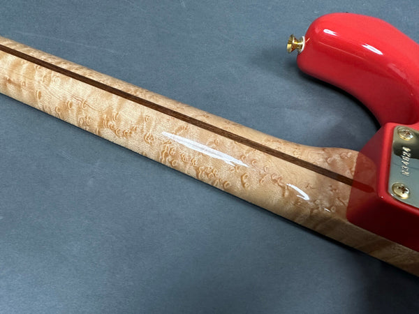 Close-up of a guitar neck made from figured maple with a contrasting dark wood stripe, showing the back of a red guitar body and strap button against a gray background.