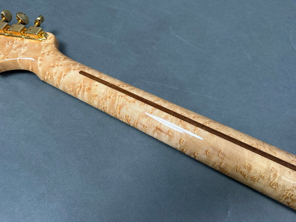 Close-up of guitar neck made from figured maple wood with visible grain pattern and gold tuning pegs on a gray background