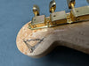 Close-up of guitar headstock with gold tuning pegs and signature logo on light-colored figured wood grain