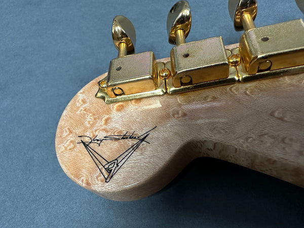 Close-up of guitar headstock with gold tuning pegs and signature logo on light-colored figured wood grain