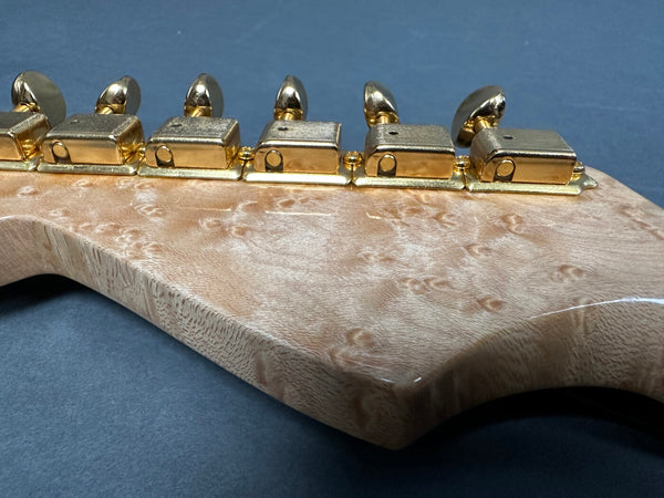 Close-up of guitar headstock with gold tuning pegs and birdseye maple wood grain finish against dark background