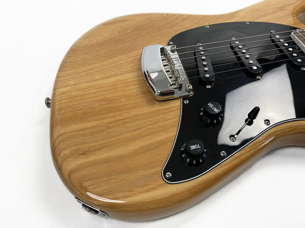 Close-up of natural wood electric guitar body with black pickguard, three single-coil pickups, volume and tone control knobs, and chrome bridge on white background