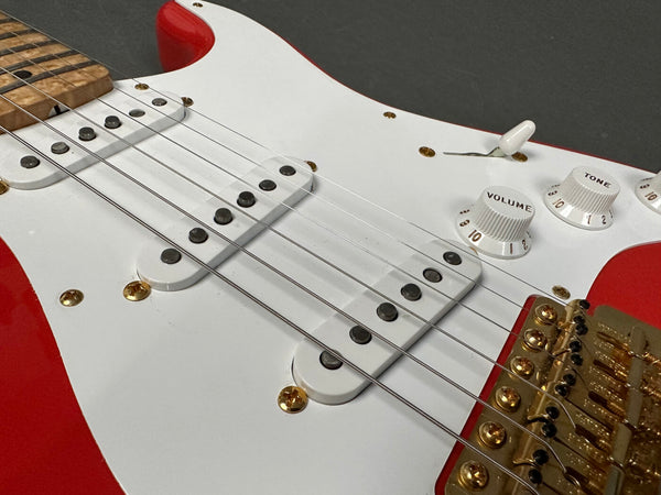 Close-up of red electric guitar body with white pickguard, maple fretboard, three single-coil pickups, white volume and tone knobs, and gold hardware bridge and screws