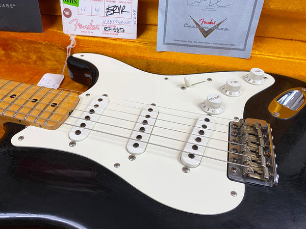 Close-up of black Fender Stratocaster electric guitar body with white pickguard, three single-coil pickups, volume and tone knobs, and vintage-style tremolo bridge inside orange guitar case with Fender Custom Shop certificate and tags visible