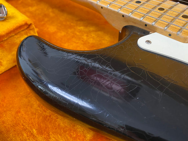 Close-up of a black electric guitar body showing aged crackled finish and a white pickguard on an orange plush-lined case background.