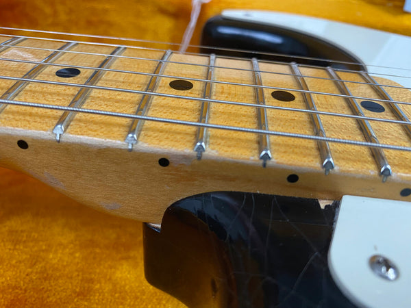 Close-up of maple guitar neck and fretboard with metal frets and black dot inlays, showing the strings and part of the black and white electric guitar body on an orange textured background.