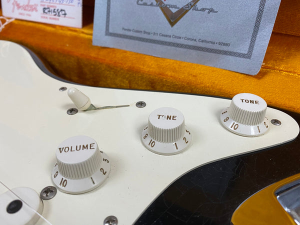 Close-up of white volume and tone control knobs on a Fender electric guitar with vintage-style cream pickguard and toggle switch, showing part of certification paper in orange-lined guitar case