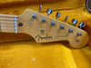 Close-up of a vintage Fender Stratocaster maple headstock with six tuning pegs, stamped with "Stratocaster" and "Original Contour Body," resting on a yellow plush guitar case lining.