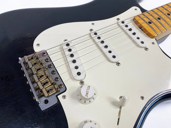 Close-up of black electric guitar body with white pickguard, three single-coil pickups, volume knob, and vintage-style bridge showing Fender branding and aged metal parts