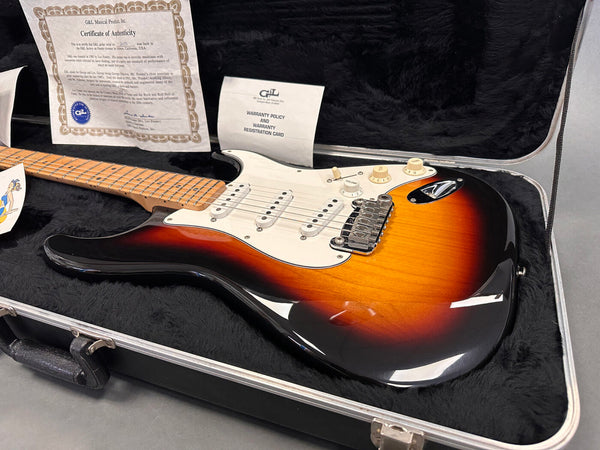 G&L electric guitar in sunburst finish resting in black plush-lined hard case with certificate of authenticity and warranty card visible