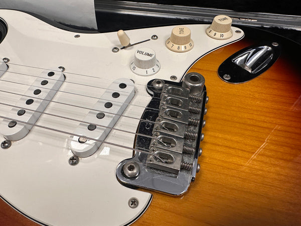 Close-up of sunburst electric guitar body showing white pickguard, three single-coil pickups, chrome bridge, and volume and tone control knobs