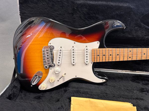 Electric guitar with sunburst finish, white pickguard, maple neck, and three single-coil pickups resting in black plush guitar case