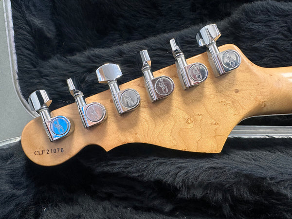 Close-up of guitar headstock showing six chrome tuning pegs with engraved logos, natural wood finish, and serial number CLF21076 on black fur background