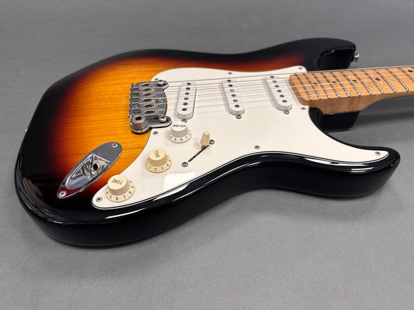 Close-up of sunburst electric guitar body with white pickguard, three single-coil pickups, and maple neck on gray background