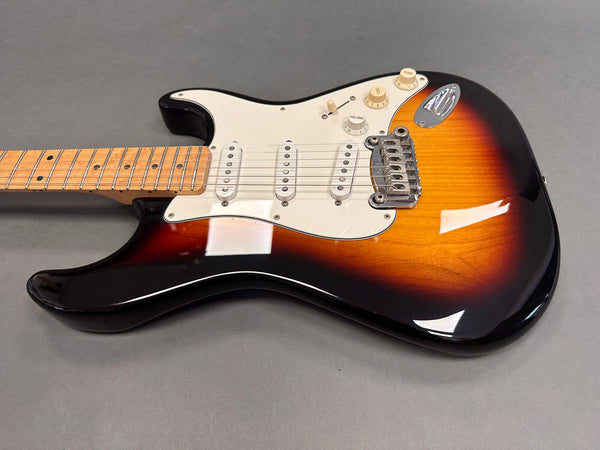 Sunburst electric guitar with maple fretboard and white pickguard resting on gray surface, close-up view of body and neck showing pickups, volume and tone knobs, and bridge.