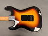 Back of sunburst electric guitar body with black plastic tremolo cavity cover and maple neck with metal neck plate on gray background