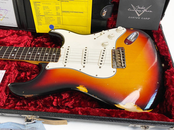 Fender Custom Shop sunburst Stratocaster electric guitar with aged body finish and white pickguard, displayed in red plush-lined hard case with Custom Care booklet and yellow hardware specification sheet.