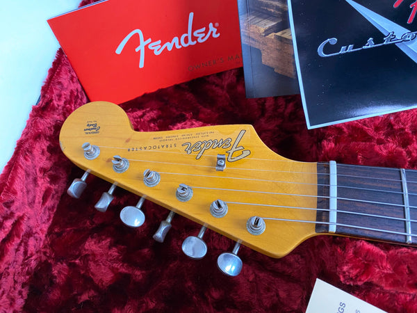 Close-up of Fender Stratocaster guitar headstock with tuning pegs and vintage logo, resting on red plush-lined guitar case with Fender owner's manual and Custom guitar booklet.