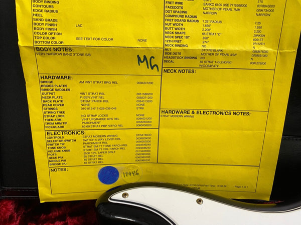 Close-up of yellow guitar specification sheet detailing vintage Strat hardware and electronics including AM vintage Strat bridge, 65 Strat pickups, and modern Strat wiring, with partial view of black and white electric guitar body.