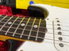 Close-up of electric guitar fretboard and strings showing white dot inlays and pickups on a white guitar body with black and yellow details on red background