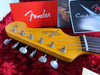 Close-up of Fender Stratocaster guitar headstock with vintage tuning pegs, showing strings and nut, with Fender owner's manual and booklet in the background on red plush fabric.