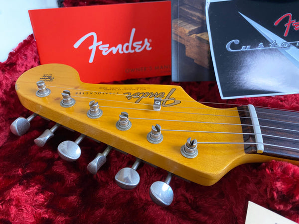 Close-up of Fender Stratocaster guitar headstock with vintage tuning pegs, showing strings and nut, with Fender owner's manual and booklet in the background on red plush fabric.