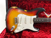 Vintage sunburst Fender Stratocaster electric guitar with white pickguard and three single-coil pickups in a red plush-lined Fender Custom case, showing worn and chipped finish.