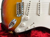 Close-up of vintage sunburst electric guitar body showing white pickguard, three single-coil pickups, volume and tone control knobs, and chrome bridge on red velvet background