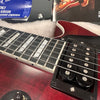 Close-up of red electric guitar body, humbucker pickups, and pearl inlay fretboard with metal strings, resting on soft white case lining with guitar magazines in background