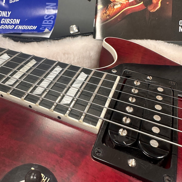 Close-up of red electric guitar body, humbucker pickups, and pearl inlay fretboard with metal strings, resting on soft white case lining with guitar magazines in background