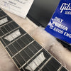 Close-up of Gibson guitar fretboard with rectangular inlays, guitar strings, and plush case lining, alongside Gibson USA blue booklet and packaging paper.