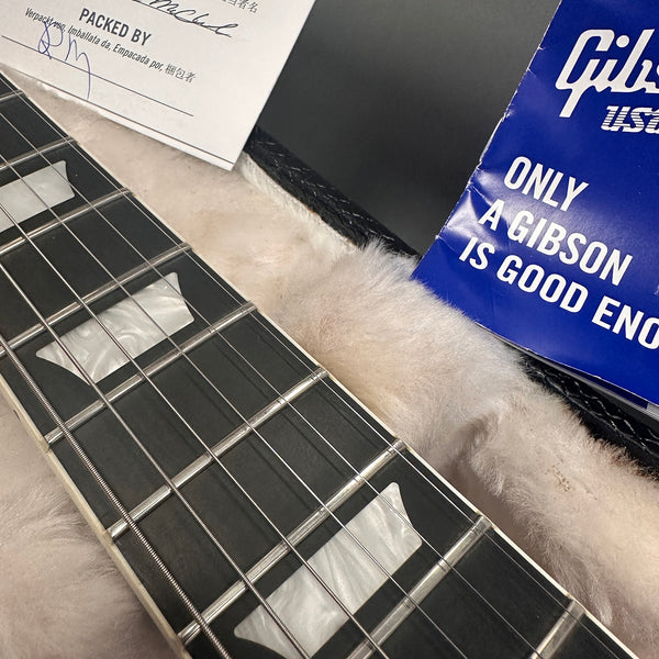 Close-up of Gibson guitar fretboard with rectangular inlays, guitar strings, and plush case lining, alongside Gibson USA blue booklet and packaging paper.