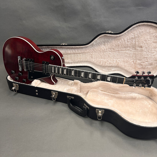 Gibson Les Paul electric guitar with dark cherry finish resting inside a black hard shell case with cream plush interior on gray background
