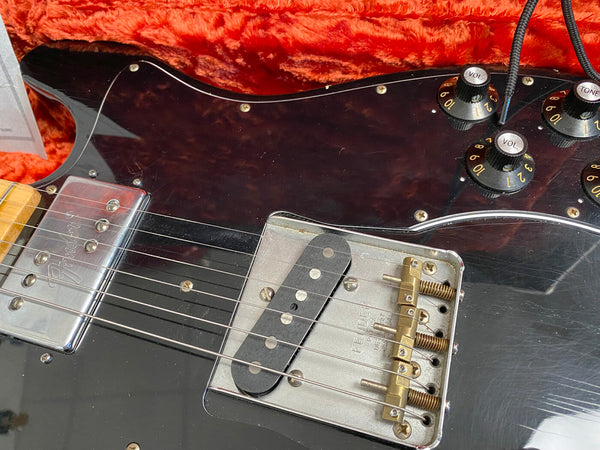 Close-up of black Fender electric guitar body showing metal bridge, single coil pickups, volume and tone control knobs, and part of the neck with strings resting on tortoiseshell pickguard
