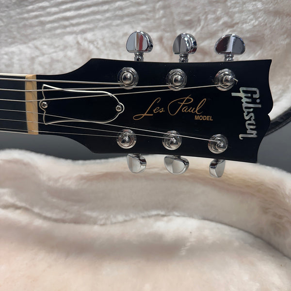 Close-up of black Gibson Les Paul Model guitar headstock with chrome tuning pegs and white binding, resting in a plush white guitar case interior.