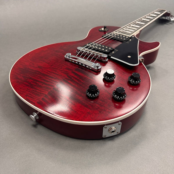 Close-up of electric guitar with red quilted maple top, black pickguard, four black control knobs, and chrome bridge on gray background
