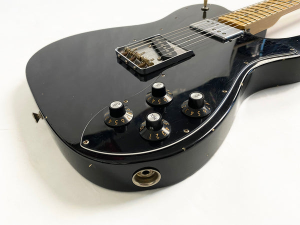 Close-up of black electric guitar body with volume and tone control knobs, metal bridge, and maple neck on white background