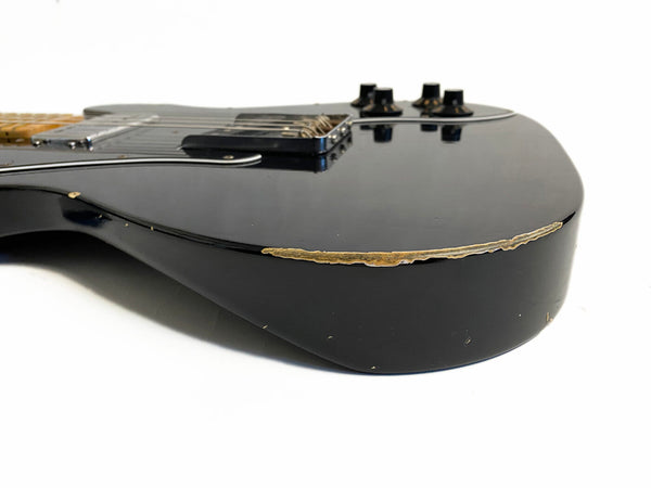 Close-up of a black electric guitar body with visible paint wear and scratches near the edge, showing vintage-style battle damage and black control knobs.