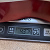 Close-up of a digital scale showing the weight of a guitar at 4.374 kilograms with the edge of a red guitar body visible on top of the scale.