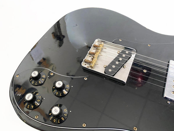 Close-up of black electric guitar body showing bridge, six strings, and four control knobs for tone and volume on a white background