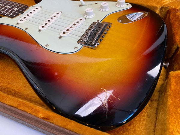 Close-up of vintage sunburst electric guitar body with white pickguard, three single-coil pickups, volume and tone control knobs in orange-lined guitar case showing signs of wear and scratches
