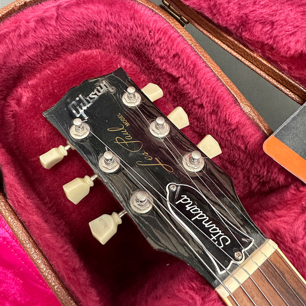 Close-up of Gibson Les Paul Standard guitar headstock with vintage tuning pegs resting in red velvet guitar case interior.